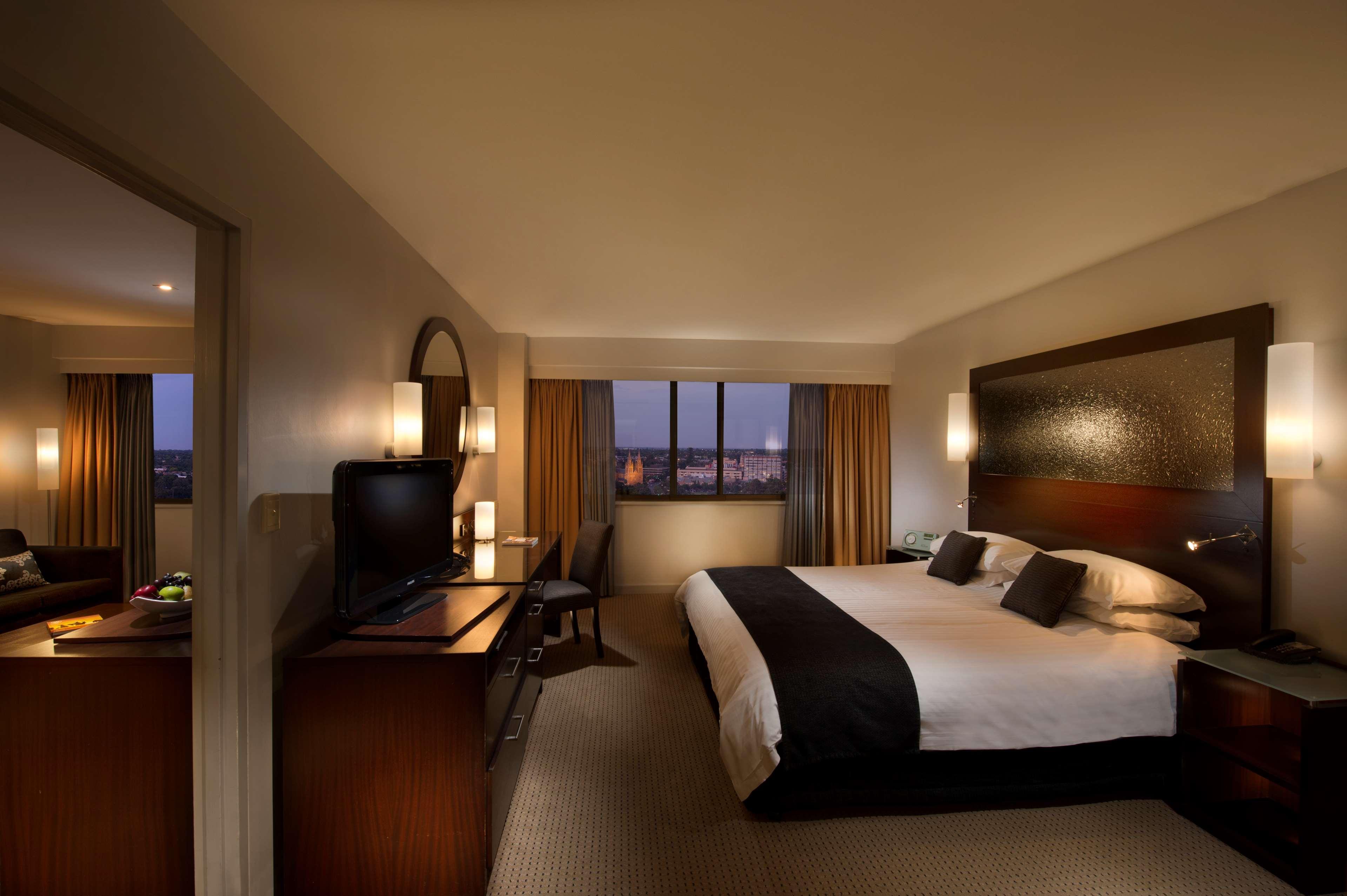 Hotel Stamford Plaza Adelaida Exterior foto The photo features a modern hotel room that includes a large bed with dark and light bedding, complemented by two pillows. The room is well-lit with warm lighting and has a contemporary aesthetic. To one side, there is a wooden dresser with a flat-sc