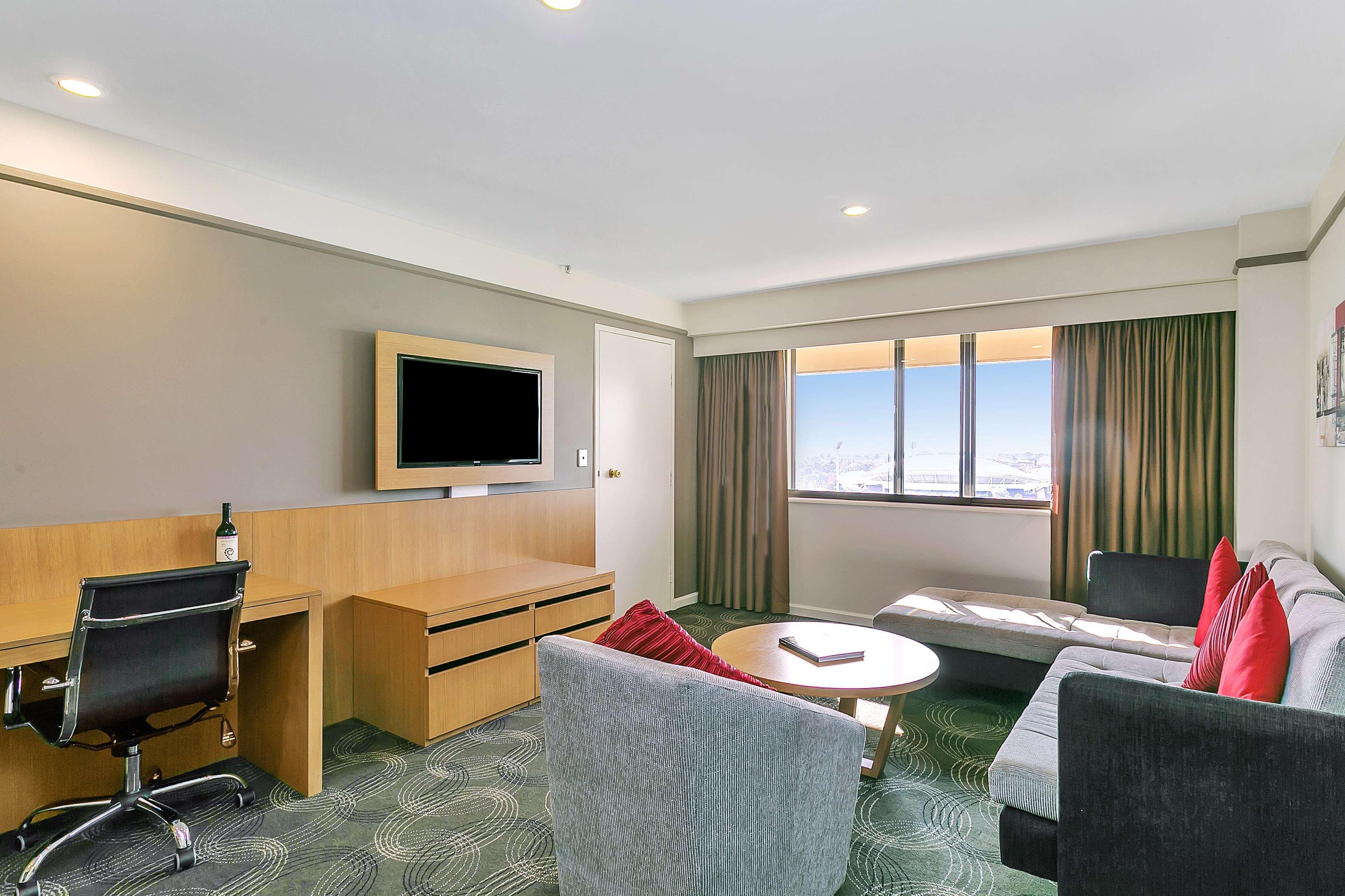 Hotel Stamford Plaza Adelaida Exterior foto The photo shows a modern hotel room or suite that features a cozy seating area. There is a comfortable sofa with red accent pillows, a round coffee table in the center, and a sleek desk with an office chair. On the wall, there is a flat-screen TV mou