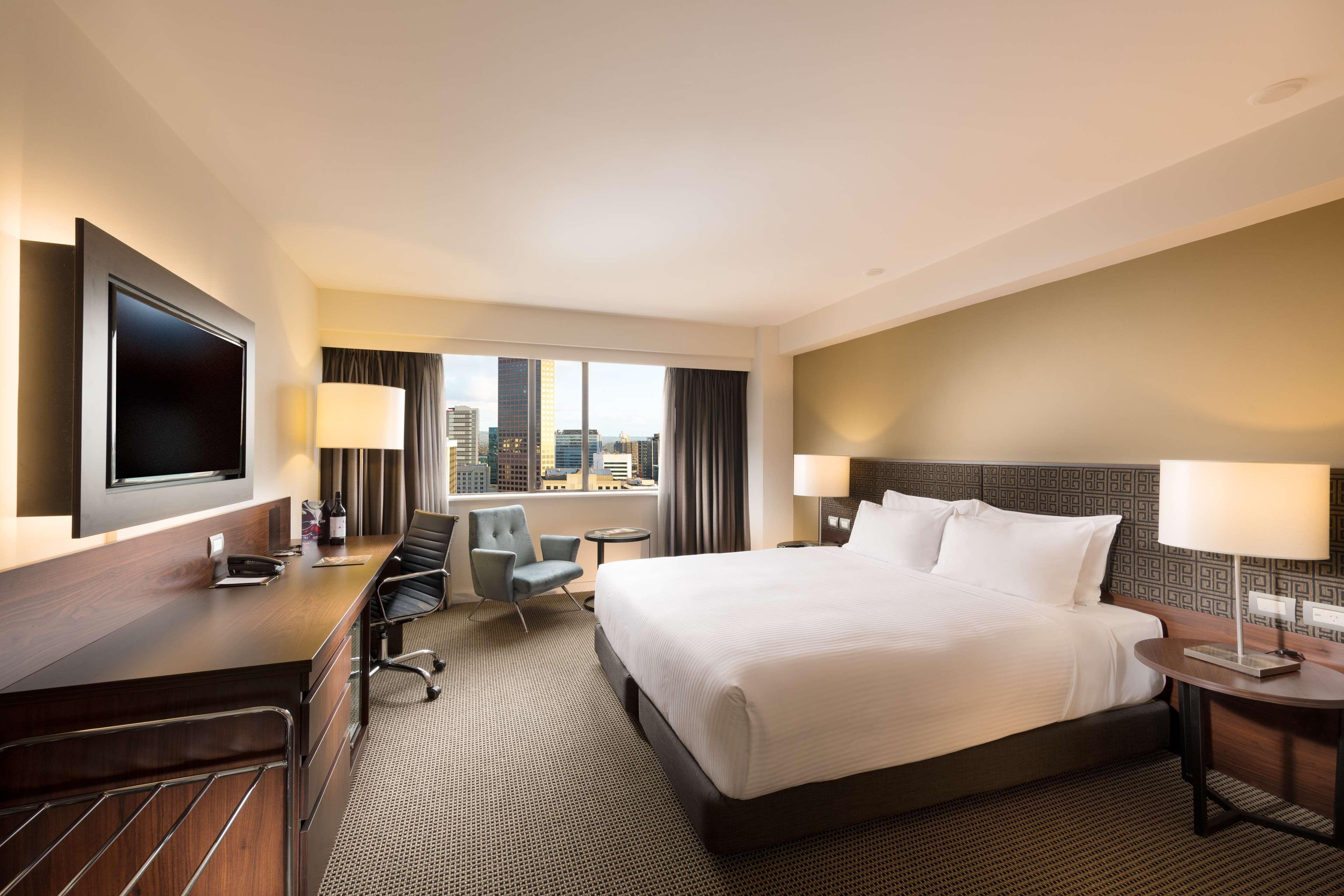 Hotel Stamford Plaza Adelaida Exterior foto The photo shows a modern hotel room with a contemporary design. It features a large bed with white linens, a bedside table on either side with lamps, and a dark wood accent wall. There's a work desk with an office chair and a flat-screen television m