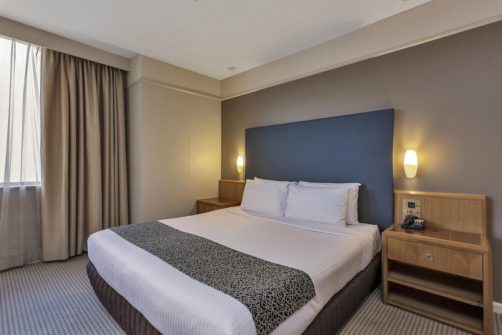 Hotel Stamford Plaza Adelaida Exterior foto The photo shows a modern hotel room. It features a double bed with a white duvet and a decorative grey and black throw. The headboard is a solid dark color. There are two bedside tables on either side of the bed, each with a lamp. Natural light enter
